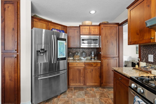 kitchen with light stone counters, appliances with stainless steel finishes, brown cabinets, range hood, and tasteful backsplash