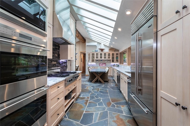 kitchen with stone tile floors, decorative backsplash, lofted ceiling with skylight, wall chimney range hood, and built in appliances