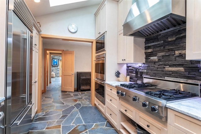 kitchen with light stone counters, stone tile floors, stainless steel appliances, backsplash, and wall chimney exhaust hood