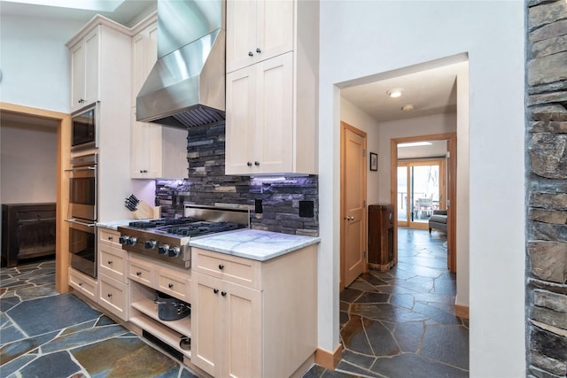 kitchen featuring stone tile floors, stainless steel appliances, backsplash, light stone countertops, and wall chimney exhaust hood