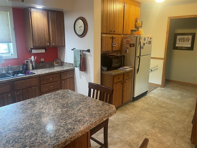 kitchen featuring black microwave, a sink, baseboards, freestanding refrigerator, and brown cabinetry