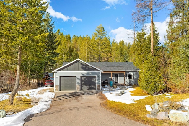 view of front facade with an attached garage, stone siding, driveway, a forest view, and board and batten siding