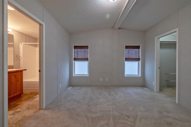 unfurnished bedroom with a textured ceiling, connected bathroom, light colored carpet, and vaulted ceiling with beams