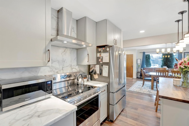 kitchen with light wood-style flooring, stainless steel appliances, wall chimney exhaust hood, tasteful backsplash, and decorative light fixtures