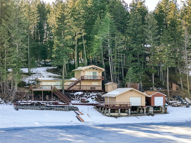 snow covered house with an outbuilding, stairway, a wooded view, and a garage