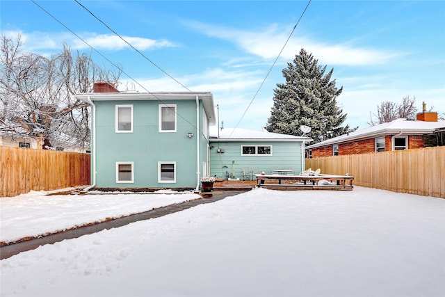 snow covered house with a deck, a chimney, fence, and stucco siding