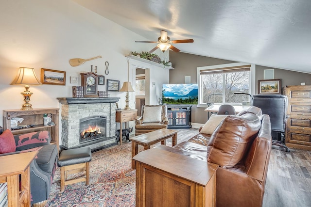 living area with ceiling fan, lofted ceiling, a stone fireplace, and wood finished floors