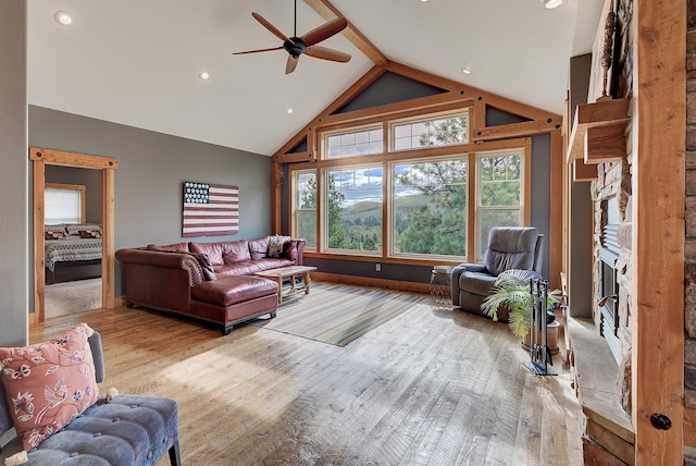 living area featuring a ceiling fan, beamed ceiling, wood finished floors, a stone fireplace, and high vaulted ceiling