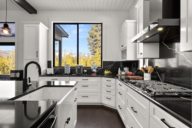 kitchen featuring dark countertops, backsplash, white cabinetry, a sink, and wall chimney range hood