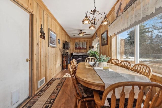 dining space with wooden walls, light wood-style flooring, ceiling fan with notable chandelier, and visible vents