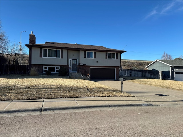 raised ranch featuring concrete driveway, a chimney, an attached garage, fence, and brick siding