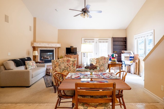 dining room with high vaulted ceiling, light colored carpet, a ceiling fan, baseboards, and a glass covered fireplace