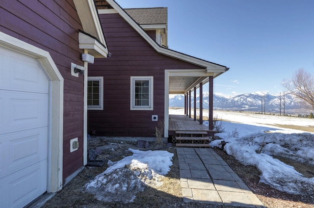 snow covered property with a porch, a mountain view, a shingled roof, and a garage