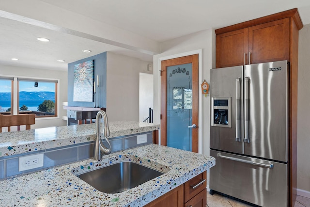 kitchen with high end fridge, a sink, recessed lighting, brown cabinetry, and light stone countertops