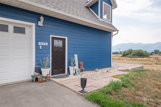 view of exterior entry with a mountain view, an attached garage, roof with shingles, and fence