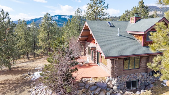 view of side of property featuring a patio, roof with shingles, log siding, a chimney, and a mountain view