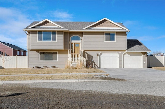 bi-level home featuring an attached garage, concrete driveway, roof with shingles, and fence