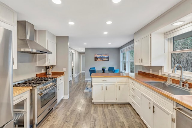 kitchen with light wood-style flooring, a sink, stainless steel appliances, a peninsula, and wall chimney range hood