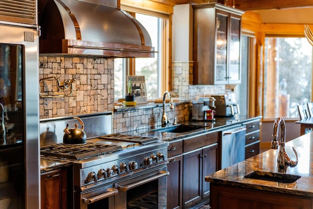 kitchen with dark stone countertops, a sink, stainless steel appliances, glass insert cabinets, and wall chimney range hood