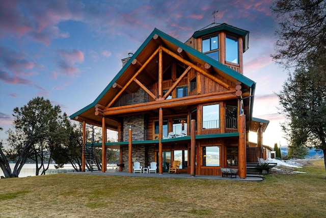 back of property at dusk with stairway, a balcony, stone siding, a patio area, and a lawn