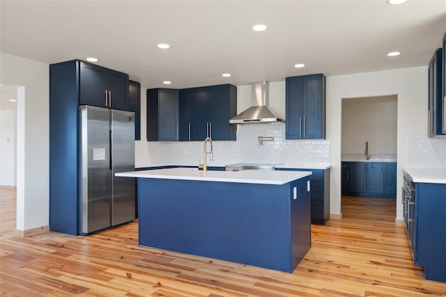 kitchen featuring stainless steel refrigerator, light wood-style flooring, blue cabinetry, and wall chimney range hood