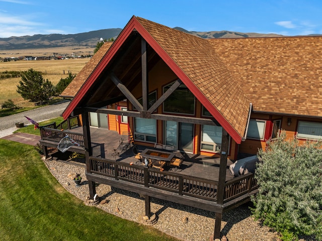 back of house featuring a lawn, a deck with mountain view, an outdoor fire pit, and a shingled roof