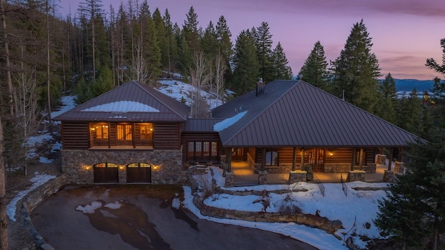 snow covered back of property featuring a standing seam roof, covered porch, a chimney, stone siding, and metal roof