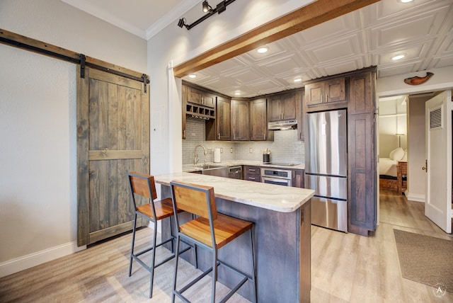 kitchen featuring under cabinet range hood, backsplash, a barn door, appliances with stainless steel finishes, and light stone countertops