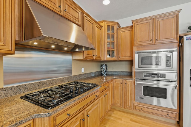 kitchen with stone counters, glass insert cabinets, under cabinet range hood, appliances with stainless steel finishes, and light wood-type flooring