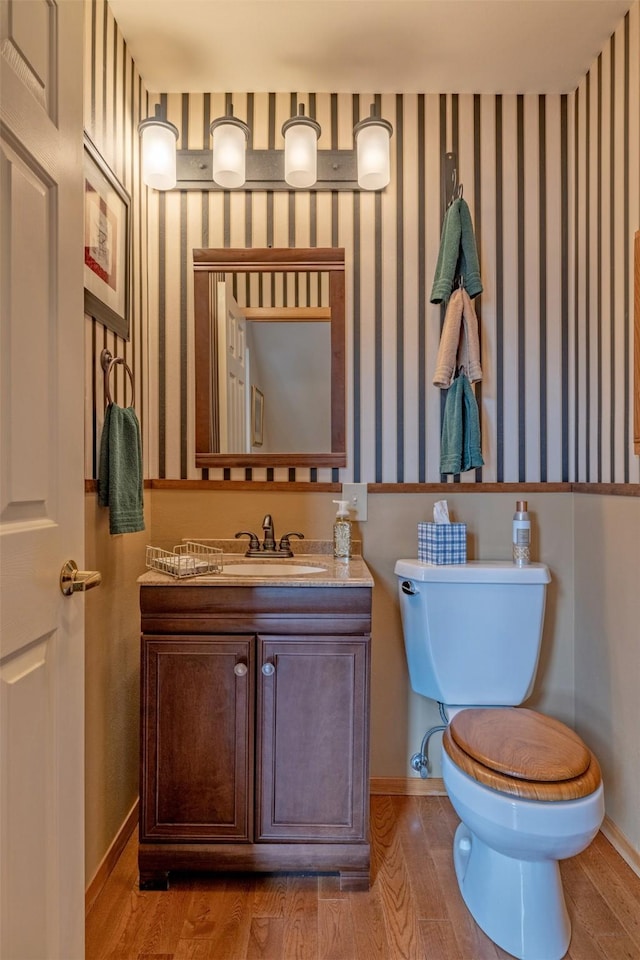 half bath featuring vanity, wood finished floors, baseboards, wallpapered walls, and toilet