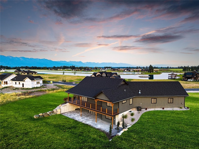 back of house at dusk featuring a lawn, a deck with mountain view, and a shingled roof