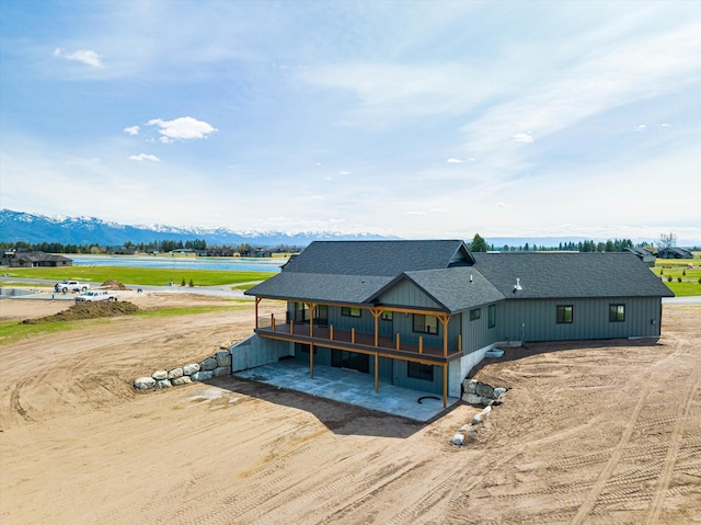 rear view of property with a shingled roof, a deck with water view, a patio, and dirt driveway