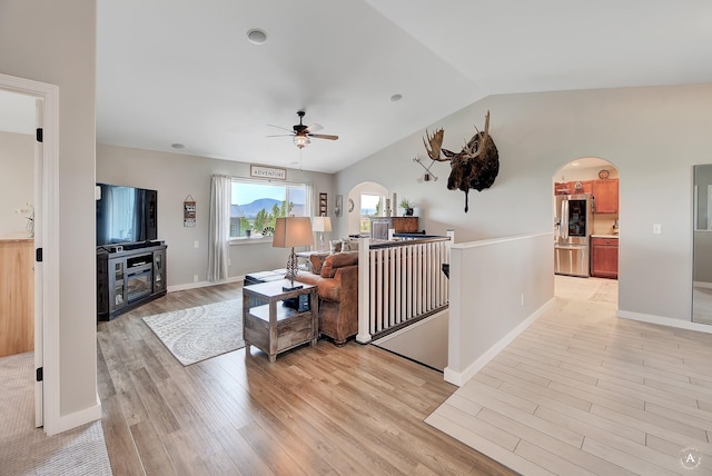 living area featuring arched walkways, light wood finished floors, baseboards, ceiling fan, and vaulted ceiling