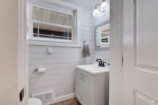 half bathroom with vanity, wooden walls, wood finished floors, visible vents, and toilet