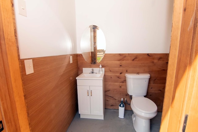 bathroom featuring wooden walls, a wainscoted wall, finished concrete floors, toilet, and vanity