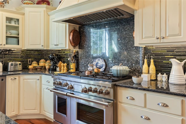 kitchen featuring custom range hood, backsplash, dark stone counters, range with two ovens, and glass insert cabinets