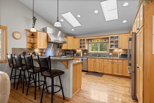 kitchen featuring stainless steel appliances, vaulted ceiling with skylight, dark stone counters, a breakfast bar area, and wall chimney exhaust hood