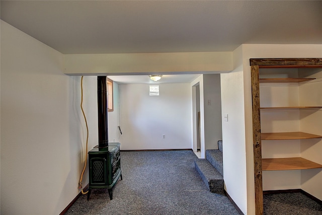 basement featuring stairs, a wood stove, baseboards, and dark carpet