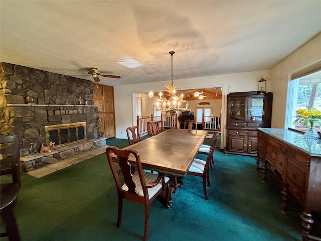 dining area with a stone fireplace, ceiling fan with notable chandelier, and dark carpet