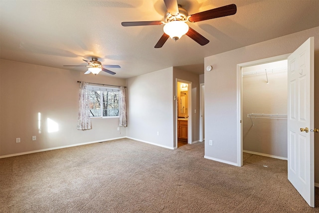 unfurnished bedroom featuring a walk in closet, ensuite bath, carpet floors, baseboards, and ceiling fan