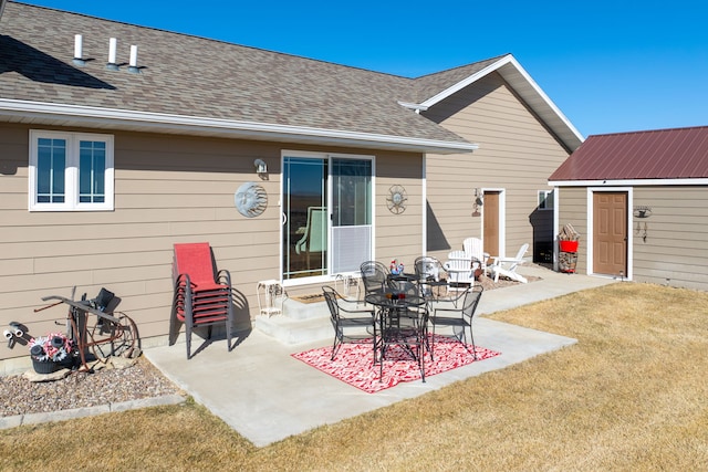 rear view of house featuring an outbuilding, a shingled roof, a lawn, and a patio area