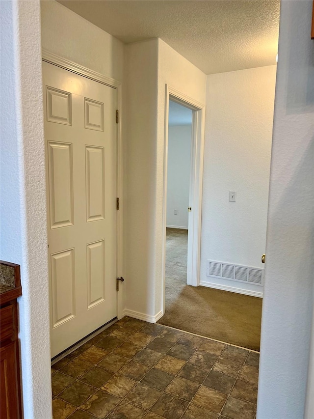 corridor featuring a textured ceiling, visible vents, stone finish flooring, baseboards, and dark colored carpet