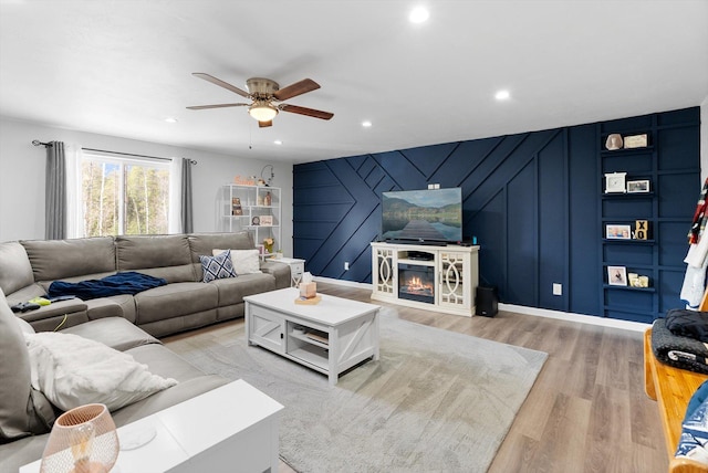 living area featuring a decorative wall, a glass covered fireplace, light wood-style flooring, a ceiling fan, and recessed lighting