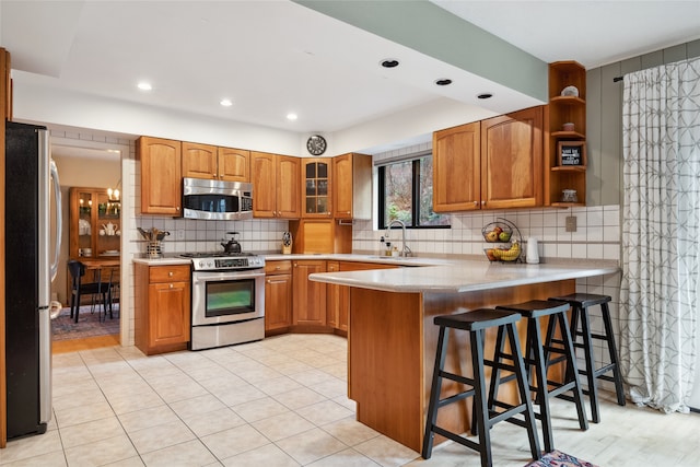 kitchen with stainless steel appliances, a sink, a peninsula, brown cabinetry, and backsplash