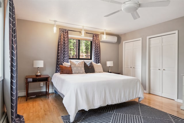 bedroom with a wall mounted air conditioner, a baseboard radiator, light wood-type flooring, two closets, and a ceiling fan