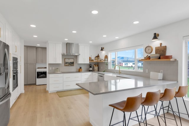 kitchen featuring stainless steel appliances, open shelves, wall chimney range hood, a sink, and a peninsula