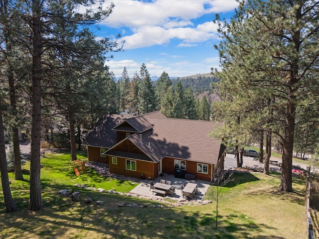 rear view of house with roof with shingles, a yard, a wooded view, and a patio