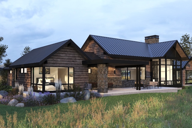 rear view of property featuring a chimney, a standing seam roof, a patio area, stone siding, and metal roof