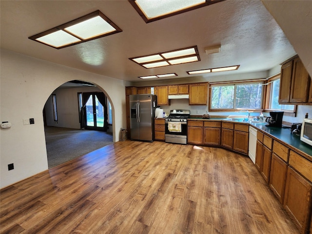 kitchen featuring under cabinet range hood, stainless steel appliances, dark countertops, arched walkways, and brown cabinetry