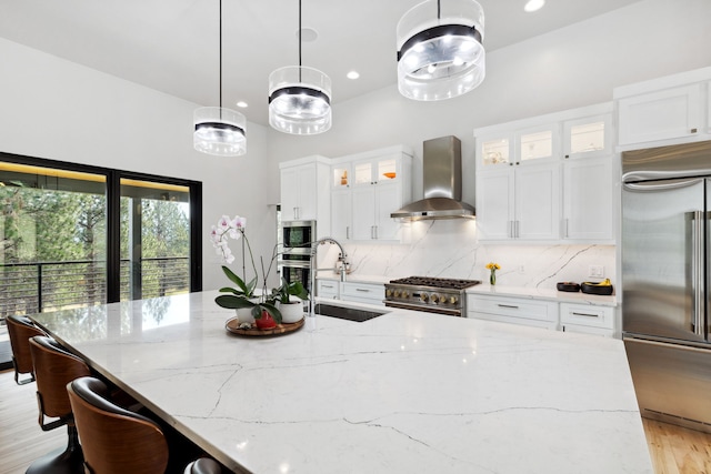 kitchen with a sink, a center island with sink, decorative backsplash, built in appliances, and wall chimney range hood